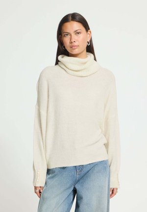 Une femme porte un pull crème à col bénitier avec un jean bleu clair et des boucles d'oreilles argentées sur un fond uni.