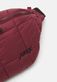 Burgundy quilted fanny pack with zip closure and black logo. Features padded texture and adjustable strap for secure wear.