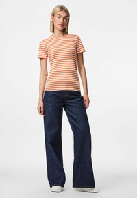 Svītraina oranža un balta īssvētku krekls ar pieguļošu formu, kas saskaņots ar tumši ziliem platājiem džinsu biksēm.