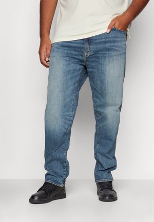 Mand iført falmede blå jeans og sorte sneakers, stående med den ene hånd i lommen mod en ensfarvet baggrund.