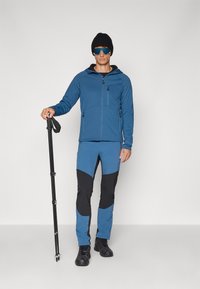 Väljasportlik riietus, mis sisaldab sinist lukuga jopet, sinise ja musta paneeliga pükse, musta beanie-mütsi ja päikeseprille. Komplektil on trekkingu kepp.