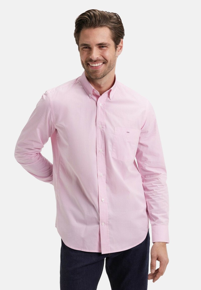 Homme souriant portant une chemise boutonnée rose pâle et un pantalon sombre, se tenant avec une main derrière le dos contre un fond uni.