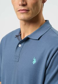 Polo tričko v tmavě modré bavlně s krátkým rukávem, s léga na dva knoflíky a tyrkysovým vyšitým logem na levé části hrudi.