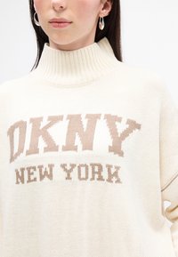 Maglione lavorato a maglia color crema con colletto alto, caratterizzato da un grande logo "DKNY" in marrone chiaro e "NEW YORK" in marrone scuro, finitura testurizzata.