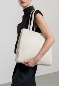 Bolso tote grande de piel beige con asas duales, textura suave y nombre de marca en relieve dorado, presentado sobre un fondo neutro.