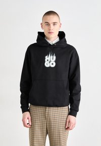 Sort hoodie med en frontlomme og et grafisk "HUGO" med flamme-accenter. Båret med beige ternede bukser. Enkel, afslappet design.