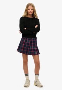 Zwarte kabelgebreide trui, geplooide marineblauwe geruite rok met rode accenten. Model draagt crèmekleurige sneakers en gestreepte sokken, met gekruiste armen.