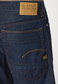 Tamsiai mėlyni džinsai G-Star Originals su odiniu ženklu, geltonais siūlais ir vienu įstrižiniu užpakaliniu kišeniu.