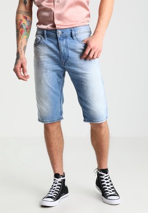 Man draagt lichtblauwe denimshorts, zwarte hoge sneakers en een lichtroze T-shirt met korte mouwen, staand met één hand in de zak.