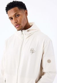Jonge man met kort, krullend haar, gekleed in een witte zip-up hoodie met beige logo borduursel op de borst en een patch op de linkermouw.