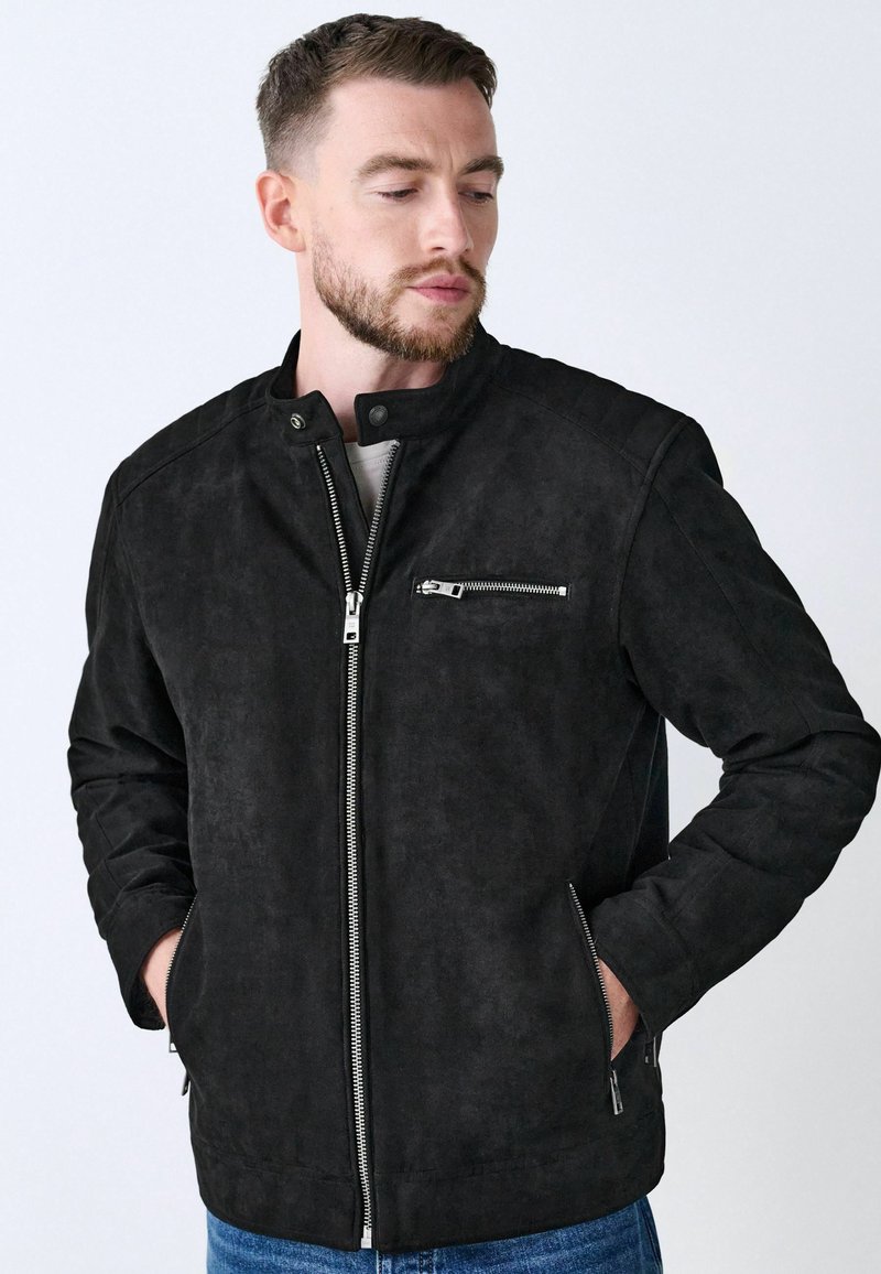 Schwarze Wildlederjacke mit Stehkragen, frontaler Reißverschluss und einer Reißverschlusstasche auf der Brust. Mit gesteppten Schulterakzenten und Seitentaschen.