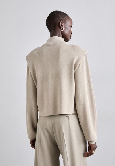 Beige korthovedet sweater med en tekstureret overflade, bred skulderdesign og ribbet kant. Sammen med lyse beige skrædderbukser.