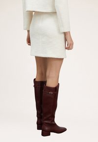 Knähöga, bordeauxfärgade läderstövlar med rundad tå, blockklack och dekorativ remdetalj nära toppen. Slät textur, elegant design.