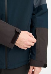 Időjárásálló dzseki, sötétkék és barna panelekből, sima textúrával, állítható tépőzáras mandzsettákkal, valamint egy biztonságos oldalzsebbel, amely húzózsinórral zárható.