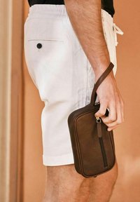 Brun läderpärm med dragkedja och handledsrem, hålls i en hand, tillsammans med vita shorts. Slät textur och rektangulär form.