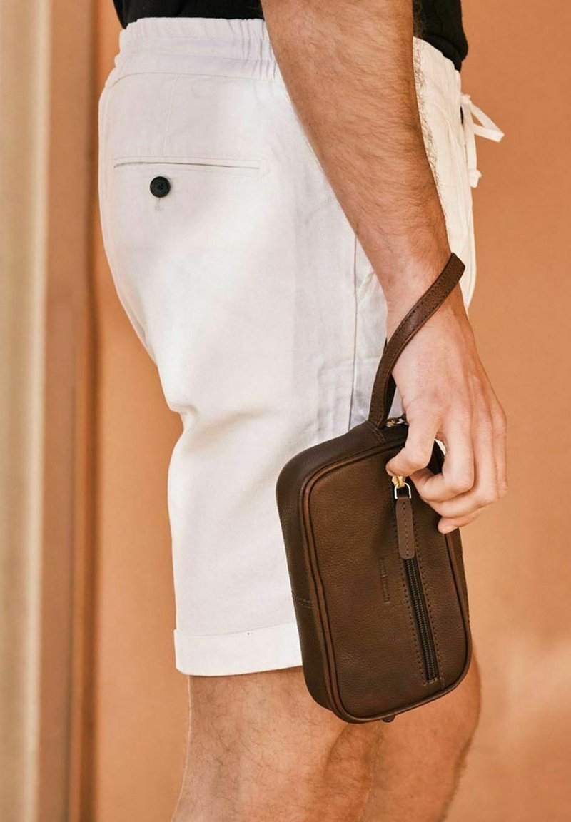 Brun läderpärm med dragkedja och handledsrem, hålls i en hand, tillsammans med vita shorts. Slät textur och rektangulär form.