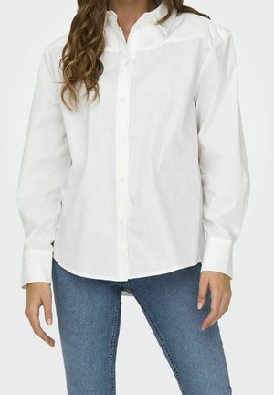 Vrouw met een wit overhemd met lange mouwen en knopen, en een blauwe spijkerbroek, met losse krullen die over één schouder vallen.