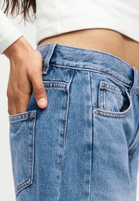 Χέρι στην πίσω τσέπη ενός φαρδιού μπλε τζιν παντελονιού που φοριέται με ένα κοντό λευκό τοπ, που δείχνει το μέσο σώμα.