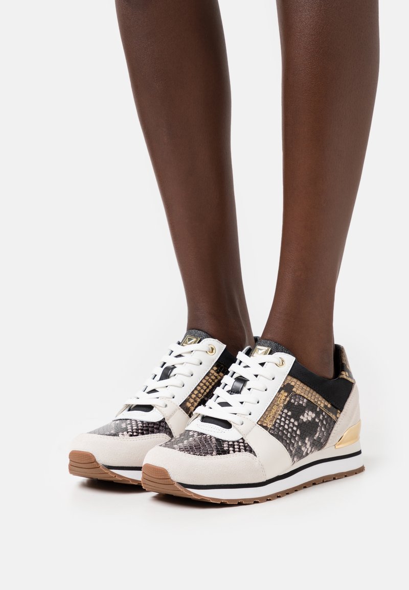 Jambes portant des baskets blanches et beiges avec un motif peau de serpent, des accents dorés, des lacets blancs et des semelles en caoutchouc marron sur fond blanc.