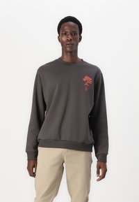 Mörkgrå sweatshirt med rund hals, ribbade ärmslut och midja, med en röd trädgrafik på vänster bröst. Slät textur och avslappnad design.