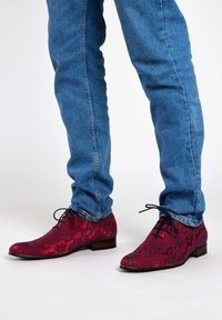 Rode, met een patroon bedekte schoenen van stof met een kantpatroon, met een spitse neus en donkere veters, gecombineerd met blauwe spijkerbroek.