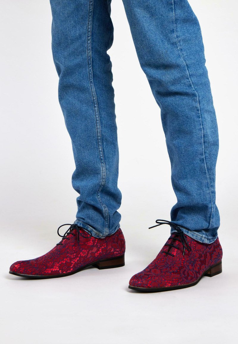 Rode, met een patroon bedekte schoenen van stof met een kantpatroon, met een spitse neus en donkere veters, gecombineerd met blauwe spijkerbroek.
