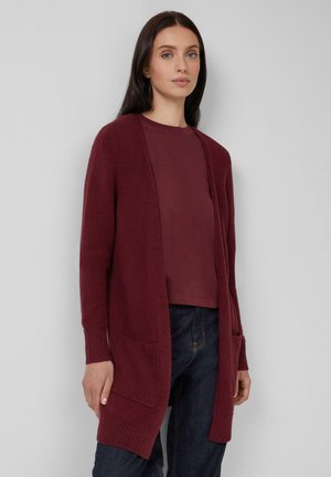 Femme portant un long cardigan bordeaux à ouverture frontale avec deux poches avant sur une chemise bordeaux assortie et un jean noir.