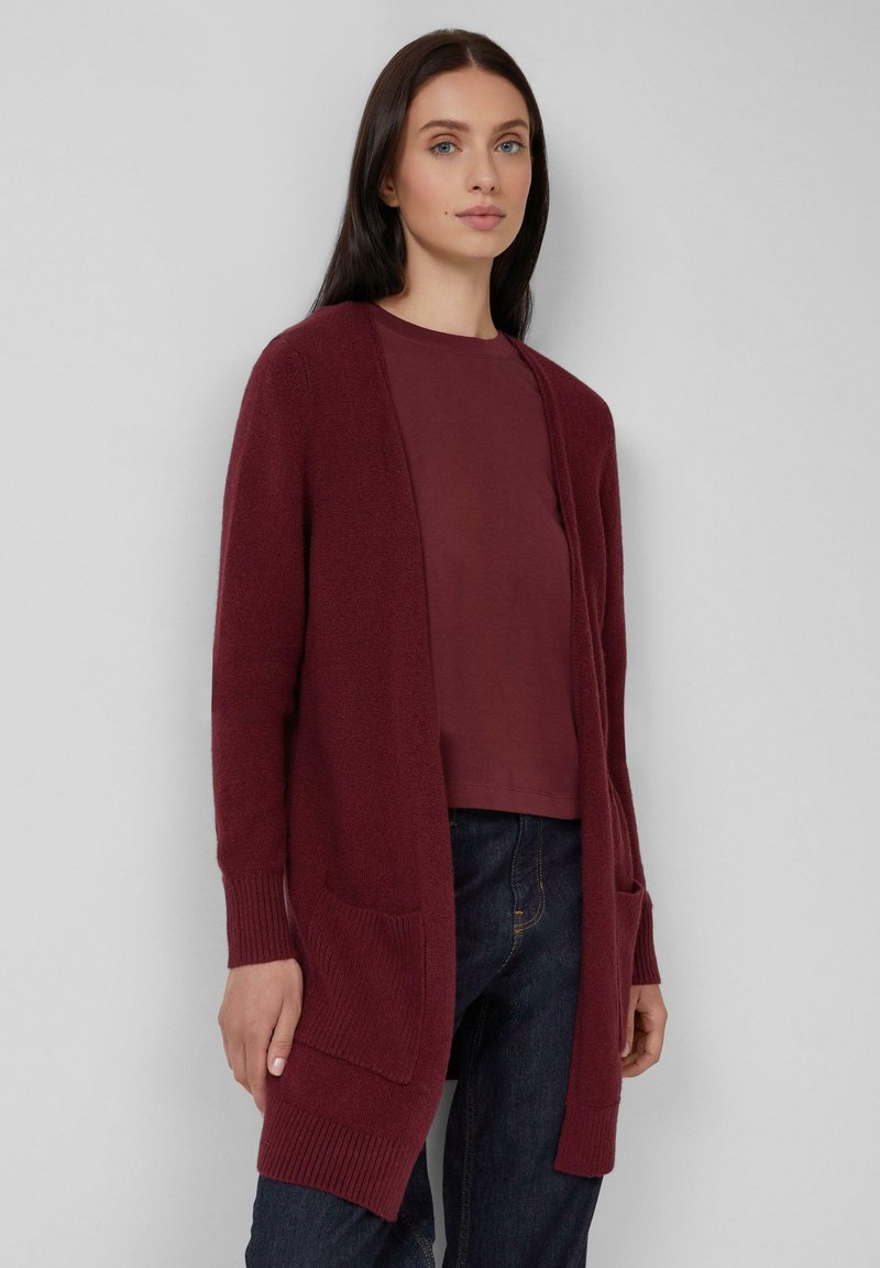 Femme portant un long cardigan bordeaux à ouverture frontale avec deux poches avant sur une chemise bordeaux assortie et un jean noir.