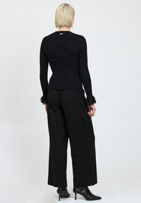 Maglione nero a maniche lunghe con dettagli in pizzo ai polsini, abbinato a pantaloni neri a gamba larga e stivaletti neri con tacco, visti da dietro.