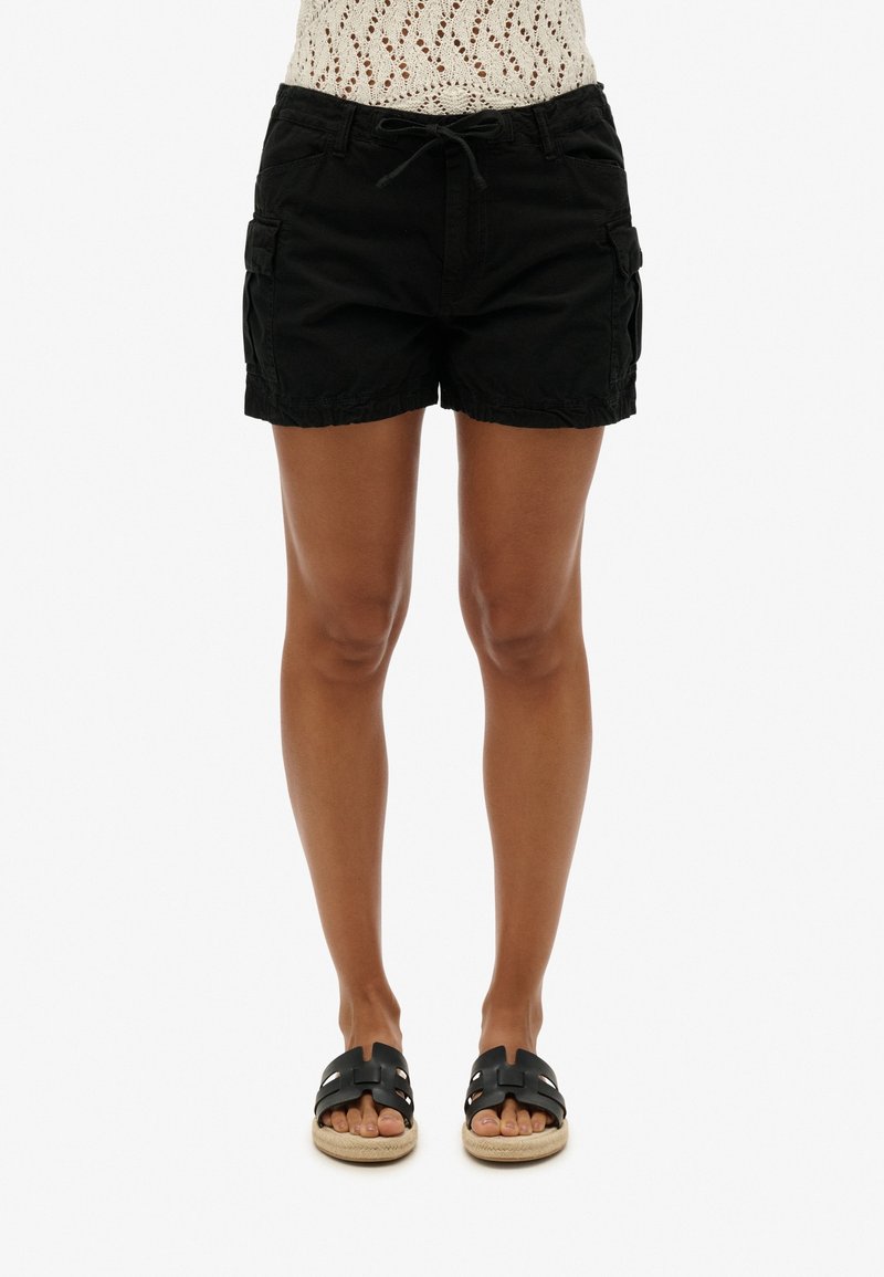 Femme portant un short cargo noir avec taille à cordon et des sandales noires à enfiler avec des semelles beige, debout devant un fond blanc.