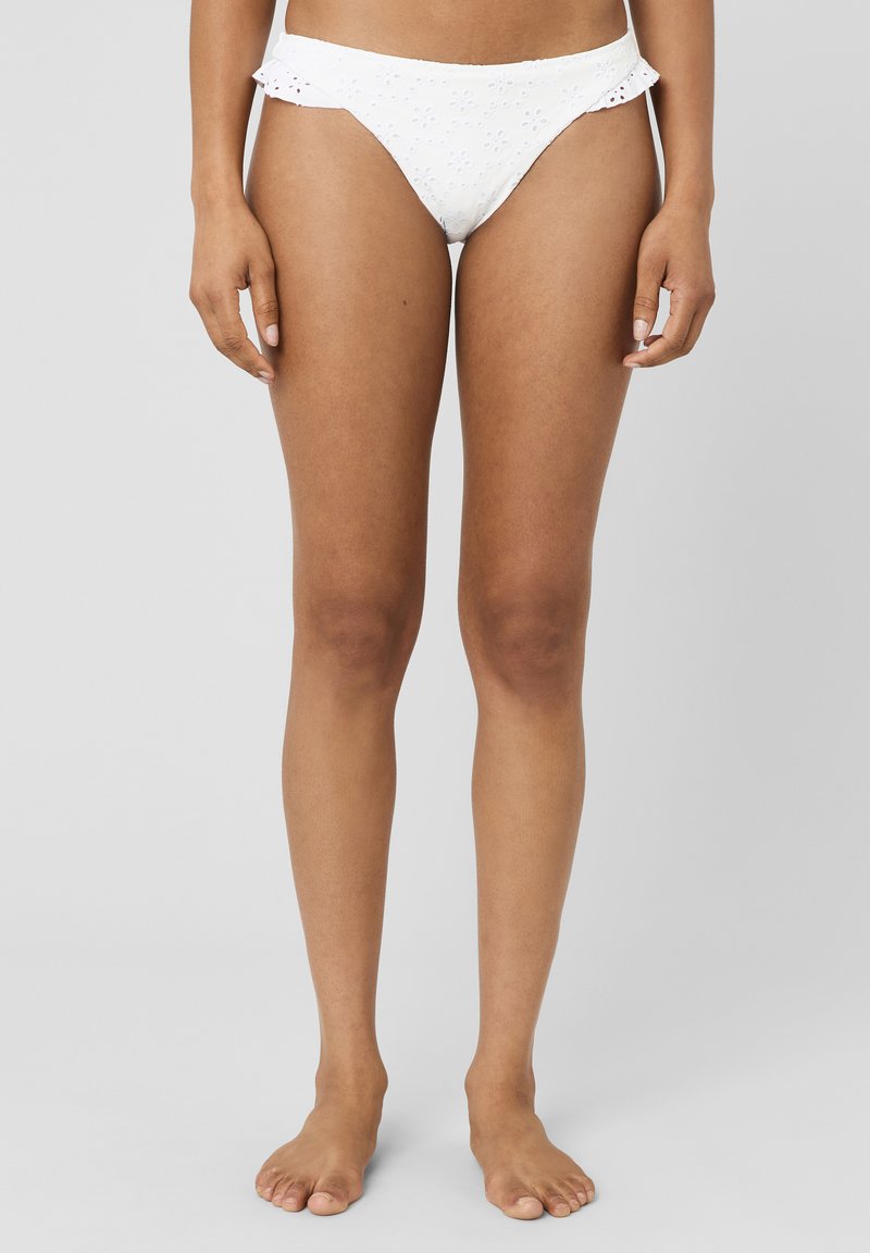 Jambes et bas du torse féminins portant un bas de bikini en dentelle fleurie blanche avec des côtés à volants, debout pieds nus sur fond gris.