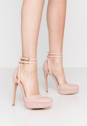 Fødder iført nude pink ruskind højhælede sko med dobbelte ankelremme og guldspænder på en hvid gulv og baggrund.