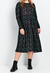 Robe noire à manches longues avec des pois teal, présentant une taille froncée et une jupe midi ; associée à des bottes noires à lacets à la cheville.
