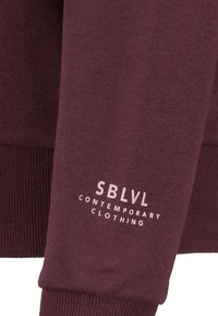 Burgundy sweatshirt with ribbed cuffs, featuring white text "SBLVL CONTEMPORARY CLOTHING" on the sleeve. Soft cotton blend material.