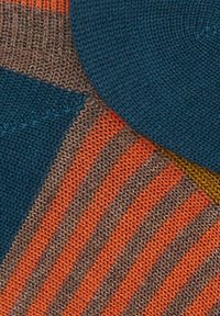 Primo piano di un tessuto lavorato a maglia con strisce blu, arancioni e marroni, e forme turchesi che formano un motivo geometrico.