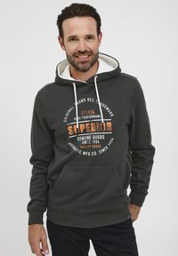Sudadera verde oscura con forro blanco en la capucha, bolsillo frontal y texto impreso en naranja y blanco. Presenta puños y dobladillo acanalados.