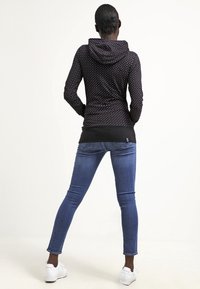 Sudadera negra con lunares blancos, con un diseño ajustado y capucha, combinada con jeans ajustados azules y zapatillas blancas.