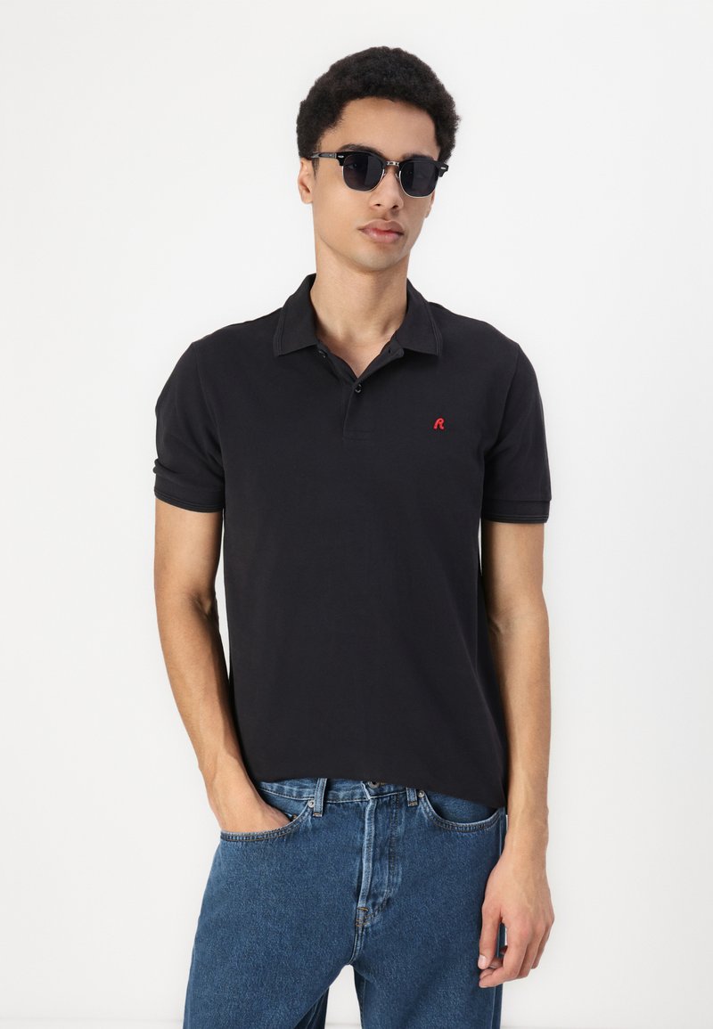 Camisa polo negra con un logo rojo en el pecho, de manga corta y con cuello. Combinada con jeans azules, complementada con gafas de sol redondas.