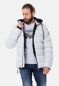Leichte graue Pufferjacke mit schwarzer Kapuze und Reißverschluss. Verfügt über ein gestepptes Design und elastische Bündchen. Darunter ein weißes Grafik-T-Shirt.