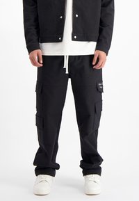 Zwarte cargobroek met meerdere zakken, een trekkoord in de taille en een gestructureerde stof. Combi met een wit t-shirt en een zwarte jas.