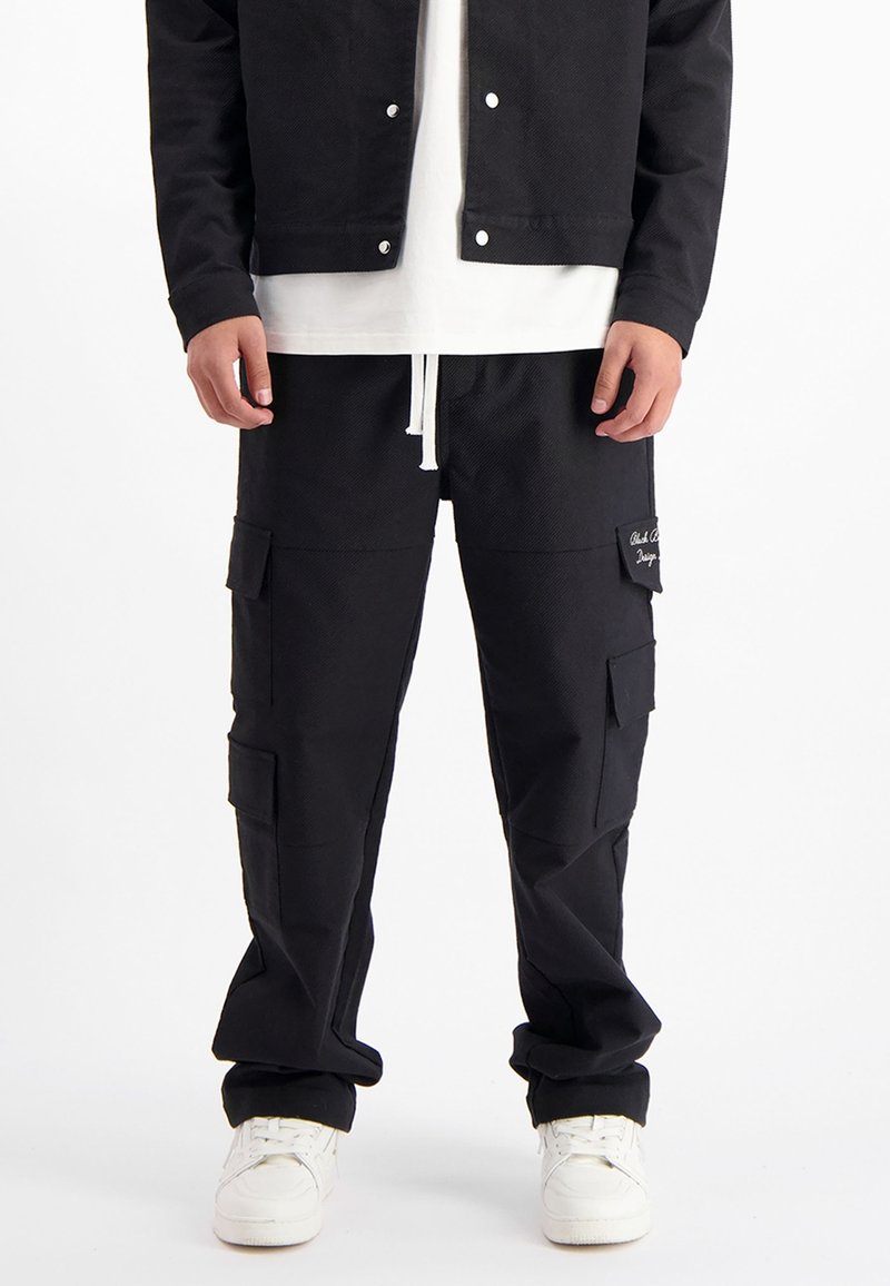 Zwarte cargobroek met meerdere zakken, een trekkoord in de taille en een gestructureerde stof. Combi met een wit t-shirt en een zwarte jas.