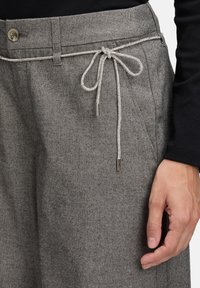 Grijze visgraatbroek met een sprankelende zilveren trekkoord dat in een strik bij de taille is gebonden, en een hand die naast de heup rust.