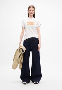 Biała koszulka z krótkim rękawem z pomarańczowym napisem "TOMMY JEANS", w połączeniu z szerokimi spodniami dżinsowymi w ciemnym kolorze i beżową kurtką opartą na ramieniu.