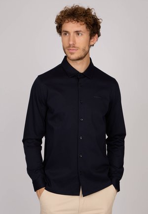 Chemise boutonnée bleu marine, avec manches longues, fabriquée dans un tissu lisse ; présente un col et un logo discret près de l'épaule.