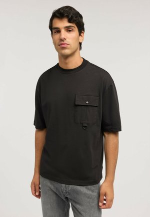 Mann trägt ein schwarzes kurzärmeliges Hemd mit einer geknöpften Brusttasche und einem Metallring, kombiniert mit grauen Jeans, steht vor einem einfarbigen Hintergrund.