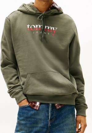 Mężczyzna ubrany w oliwkowo-zieloną bluzę Tommy Jeans z kapturem, nałożoną na czerwoną kratowaną koszulę, w niebieskich dżinsach, z rękami w kieszeniach, na białym tle.