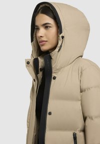 Beige pufferjack met een grote capuchon, zwarte accenten en een ritssluiting. Beschikt over drukknopen en een gewatteerde textuur voor isolatie.