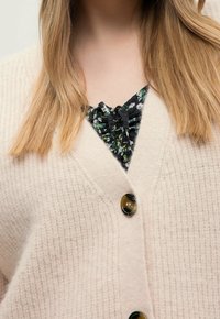 Dlouhé blond vlasy přes béžový cardigan s knoflíky a texturovaným pletením, odhalující černý květinový top pod ním s nařaseným výstřihem.