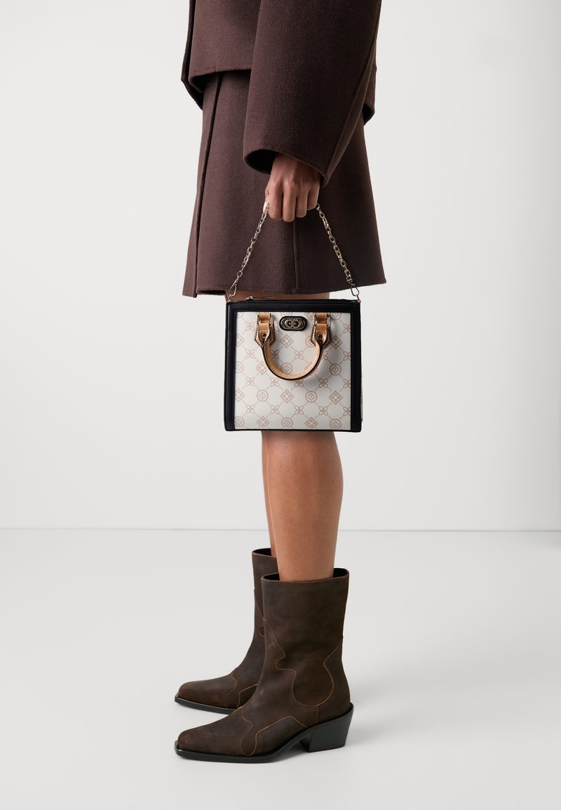Personne en manteau marron et bottes marron foncé tenant un petit sac à main carré avec un devant beige à motifs et des anses couleur tabac.