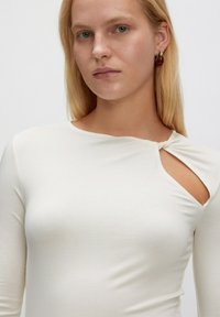 Langärmliges Creme-Oberteil mit einem verdrehten Knotendetail an der Schulter und einem asymmetrischen Ausschnitt. Hergestellt aus glattem, elastischem Stoff.